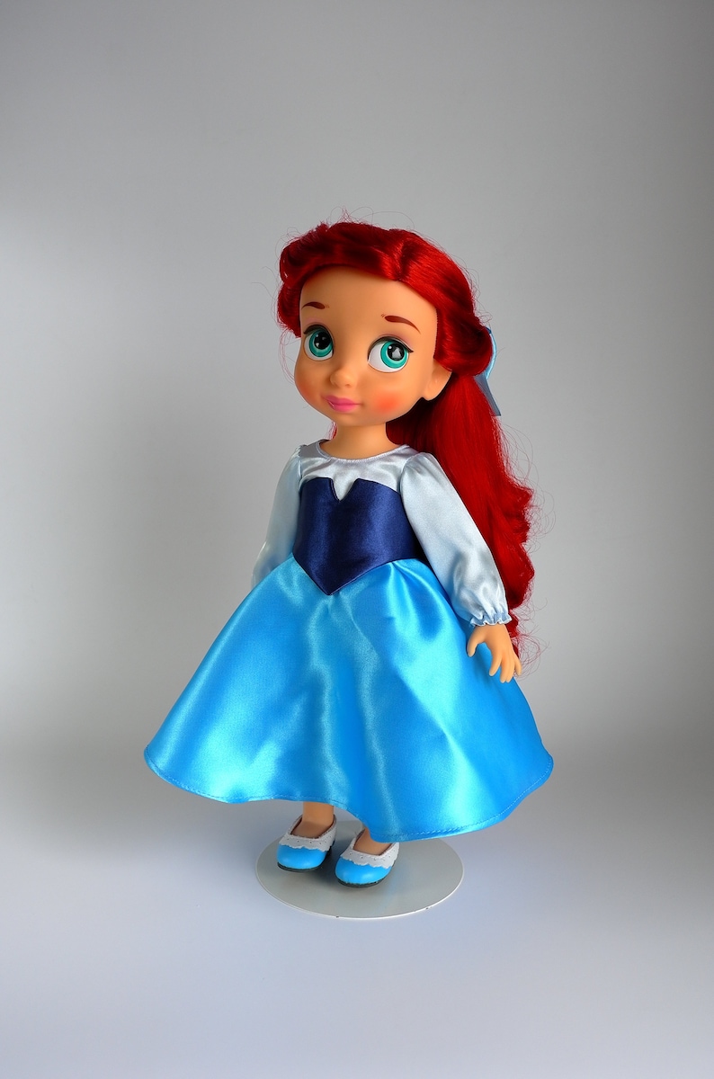 May include: A red-haired doll wearing a blue and white dress with blue shoes. The doll has a light blue top and a dark blue bodice. The doll is standing on a white stand.