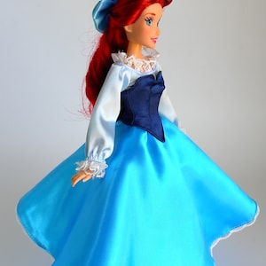 May include: A red-haired doll wearing a blue and white dress with a lace trim. The doll has a blue bodice and a long, flowing skirt.