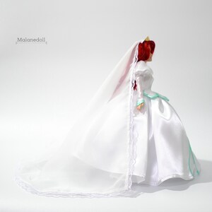 May include: A white wedding dress with a long train and a veil. The dress has a fitted bodice and a full skirt. The doll is wearing a crown.