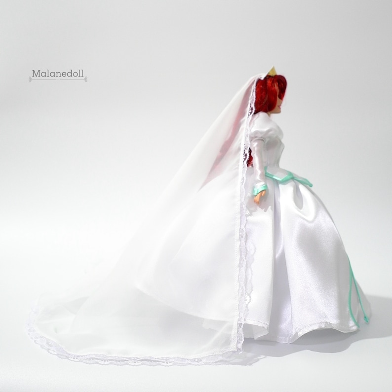 May include: A white wedding dress with a long train and a veil. The dress has a fitted bodice and a full skirt. The doll is wearing a crown.