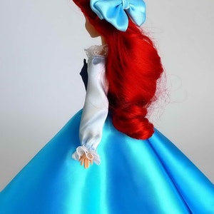 May include: A red-haired doll wearing a blue satin dress with a white lace trim. The doll has a blue bow in her hair.