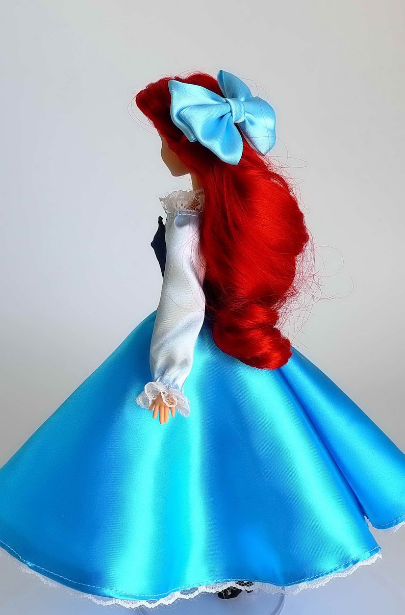 May include: A red-haired doll wearing a blue satin dress with a white lace trim. The doll has a blue bow in her hair.