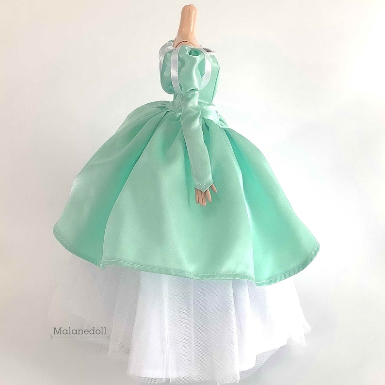 Puede incluir: Un vestido de bola de sat&eacute;n verde menta con una falda interior de tul blanco. El vestido tiene mangas abullonadas con ribete de cinta de sat&eacute;n blanco. El vestido est&aacute; en un maniqu&iacute; de tama&ntilde;o de mu&ntilde;eca.