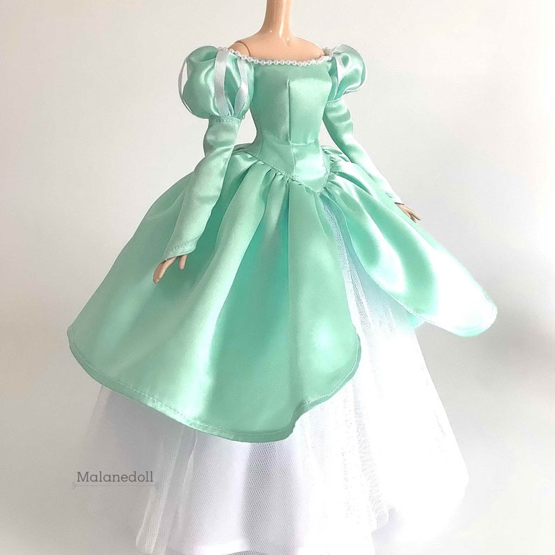 Puede incluir: Un vestido de bola de sat&eacute;n verde menta con una falda de tul blanca. El vestido tiene un escote de coraz&oacute;n y mangas abullonadas con lazos de sat&eacute;n blanco. El vestido est&aacute; dise&ntilde;ado para una mu&ntilde;eca.