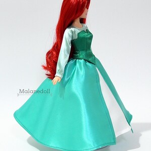 May include: A red-haired doll wearing a green and white dress. The dress has a green bodice and a white underskirt. The doll is standing on a white surface.