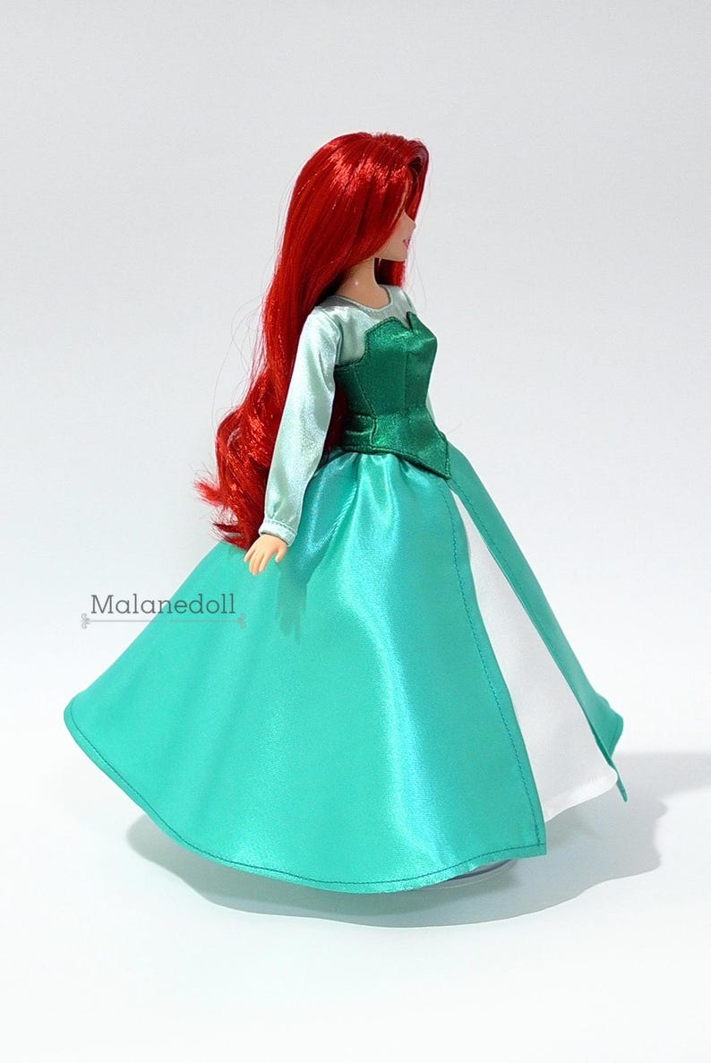 May include: A red-haired doll wearing a green and white dress. The dress has a green bodice and a white underskirt. The doll is standing on a white surface.