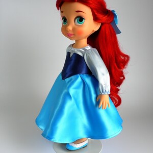 May include: A red-haired doll wearing a blue and white dress with a navy blue bodice. The doll has blue eyes and is wearing blue shoes.
