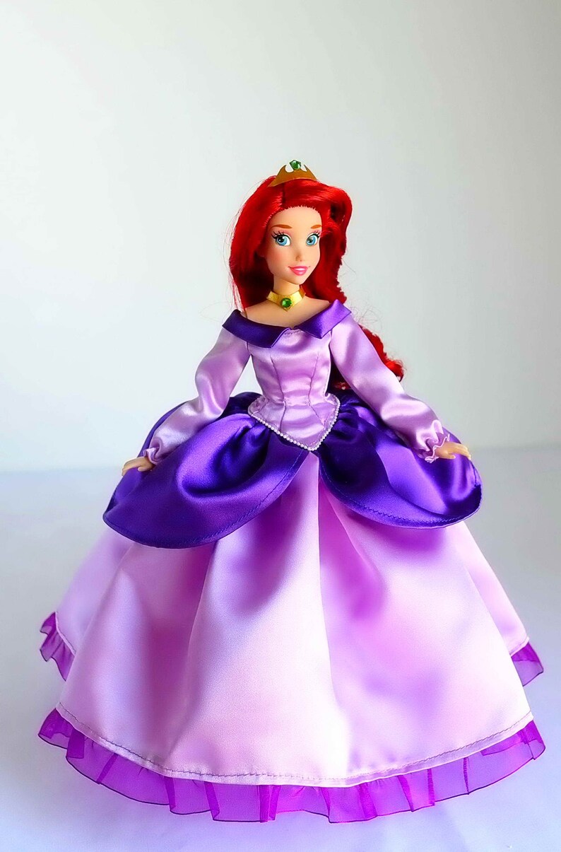May include: A red-haired doll wearing a purple and pink satin dress with a gold crown. The doll has blue eyes and is wearing a green necklace.