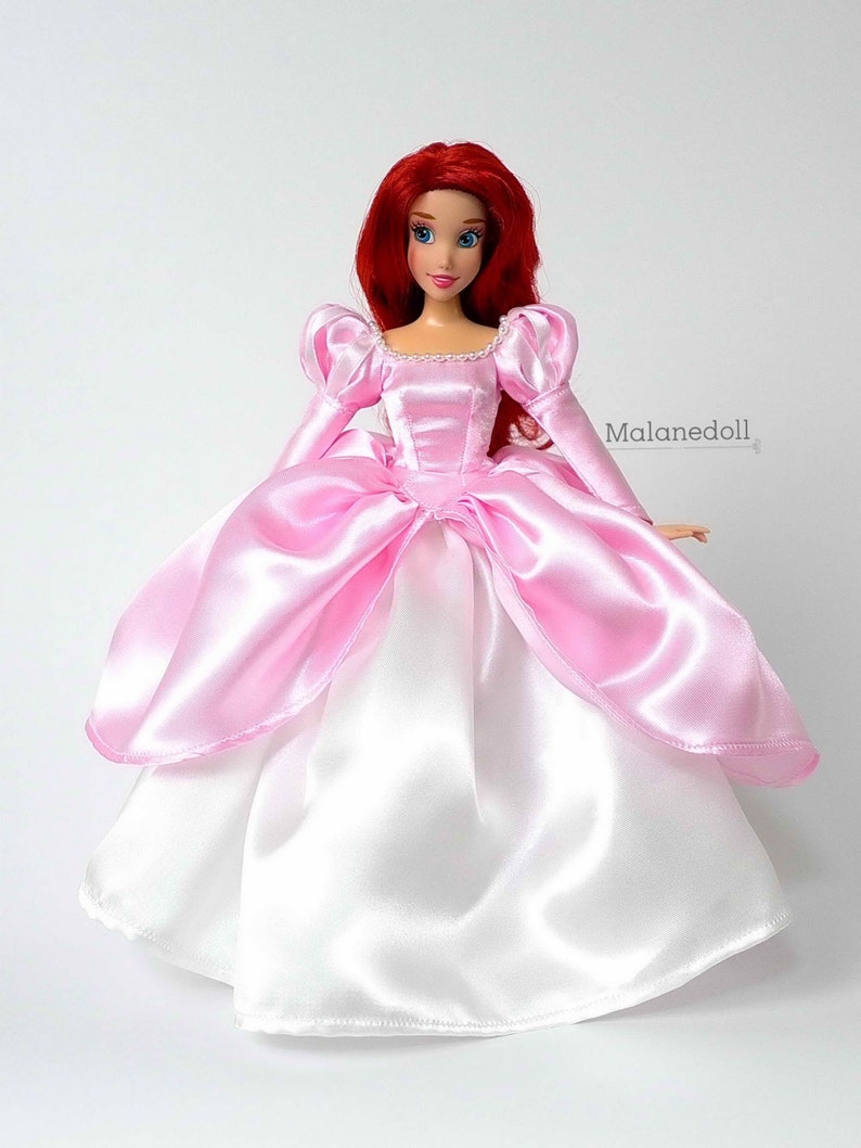 Peut inclure: Poup&eacute;e aux cheveux roux et aux yeux bleus v&ecirc;tue d'une robe de satin rose et blanche. La robe pr&eacute;sente un corsage rose avec des manches longues et une jupe ample. La poup&eacute;e est un objet de collection.