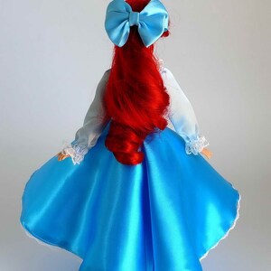 May include: A red-haired doll wearing a blue satin dress with white lace trim and a light blue bow in her hair.