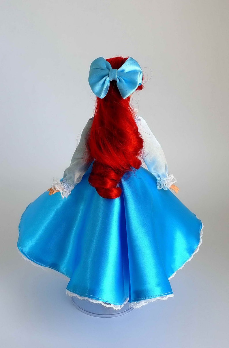 May include: A red-haired doll wearing a blue satin dress with white lace trim and a light blue bow in her hair.