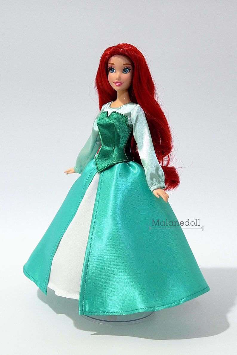 May include: A red-haired doll wearing a green and white dress. The doll has blue eyes and a red wig. The dress has a green bodice and a white skirt. The doll is standing on a clear stand.