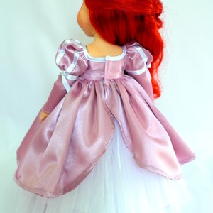 May include: A pink satin dress with white tulle underskirt and long sleeves. The dress has a white trim around the neckline and sleeves. The doll is wearing a crown.