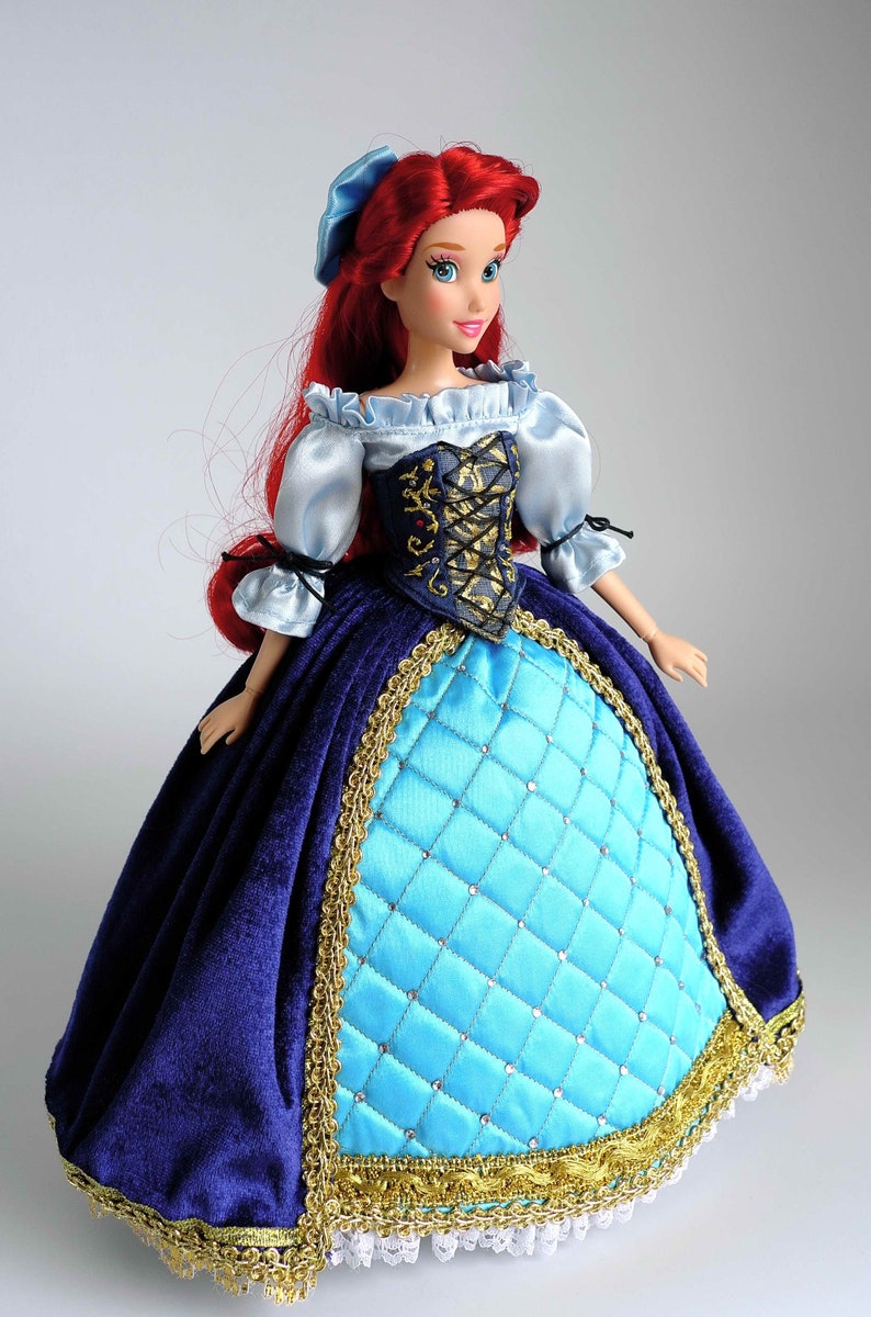 May include: A red-haired doll wearing a blue and gold dress with a white lace trim. The doll has blue eyes and is wearing a blue ribbon in her hair.