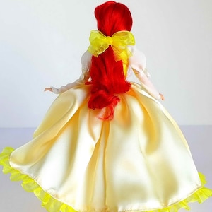 Pode incluir: Uma boneca de cabelo ruivo vestindo um vestido de cetim amarelo com uma babosa de tule amarelo na bainha. A boneca tem um la&ccedil;o amarelo no cabelo.