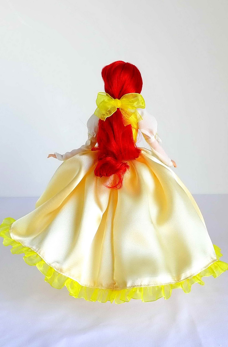 Pode incluir: Uma boneca de cabelo ruivo vestindo um vestido de cetim amarelo com uma babosa de tule amarelo na bainha. A boneca tem um la&ccedil;o amarelo no cabelo.