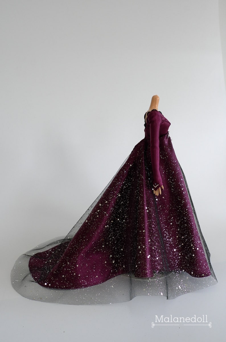 Puede incluir: Un vestido largo de color burdeos con una superposici&oacute;n de tul negro y purpurina plateada. El vestido tiene mangas largas y un corpi&ntilde;o ajustado.