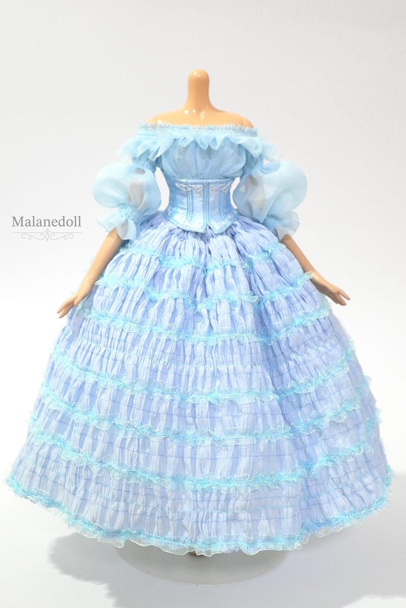 K&ouml;nnte beinhalten: Hellblaues Puppenkleid mit schulterfreiem Ausschnitt und ger&uuml;schten &Auml;rmeln. Das Kleid hat ein figurbetontes Oberteil und einen vollen, mehrlagigen Rock mit zarten Verzierungen. Das Puppenkleid ist im Vintage-Stil gehalten.