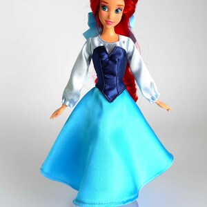 K&ouml;nnte beinhalten: Puppe einer Prinzessin mit rotem Haar, blauen Augen und hellem Teint. Die Puppe tr&auml;gt ein blau-wei&szlig;es Kleid mit einem dunkelblauen Oberteil und einer hellblauen Schleife. Die Puppe steht auf einem durchsichtigen Kunststoffst&auml;nder.