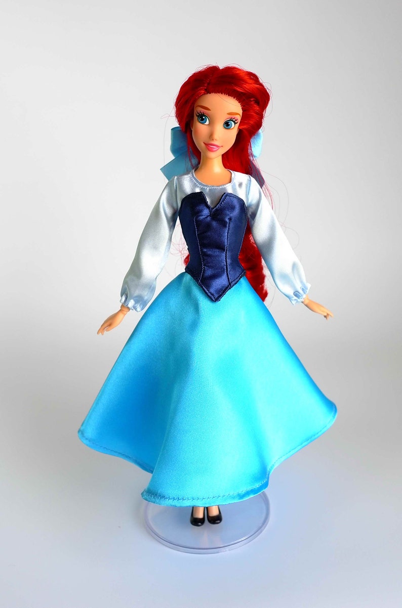 K&ouml;nnte beinhalten: Puppe einer Prinzessin mit rotem Haar, blauen Augen und hellem Teint. Die Puppe tr&auml;gt ein blau-wei&szlig;es Kleid mit einem dunkelblauen Oberteil und einer hellblauen Schleife. Die Puppe steht auf einem durchsichtigen Kunststoffst&auml;nder.