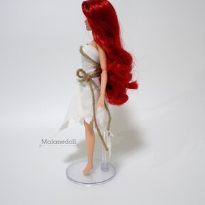 May include: A red-haired doll wearing a white dress with a brown rope belt. The doll is standing on a clear plastic stand.