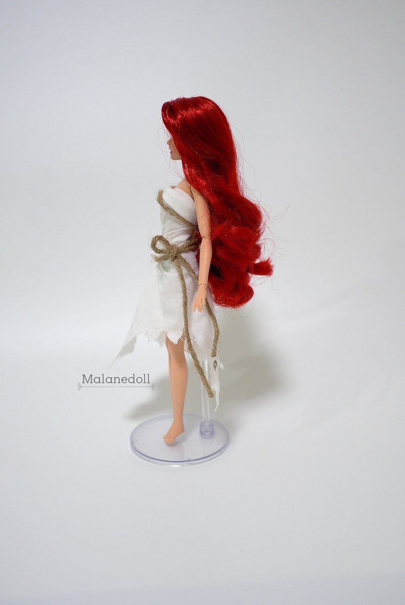 May include: A red-haired doll wearing a white dress with a brown rope belt. The doll is standing on a clear plastic stand.