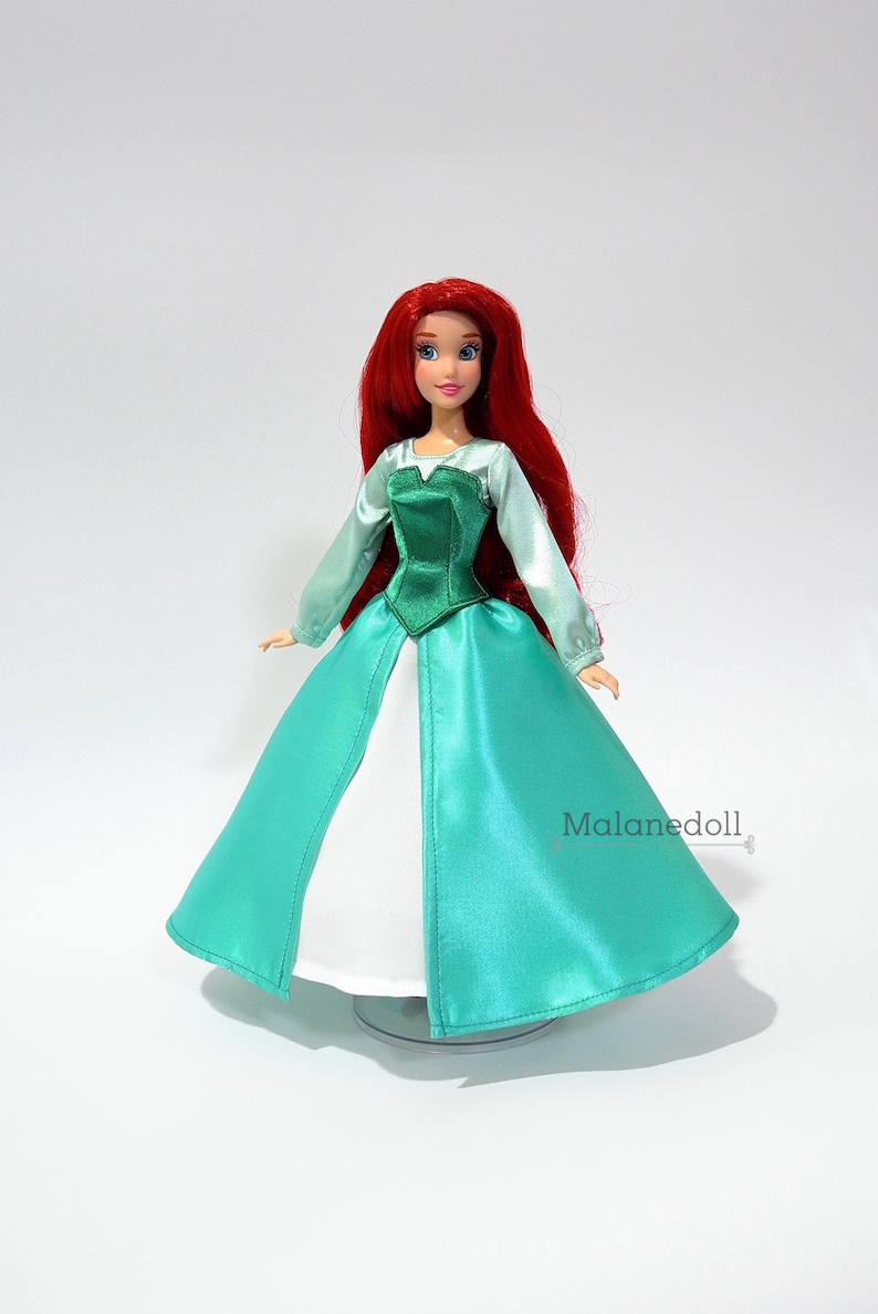 May include: A red-haired doll wearing a teal and green dress with a white underskirt. The doll has blue eyes and is wearing a crown.