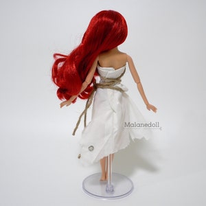 May include: A red-haired doll wearing a white dress with a rope belt. The doll is standing on a clear plastic stand. The text "Malanedoll" is visible on the dress.