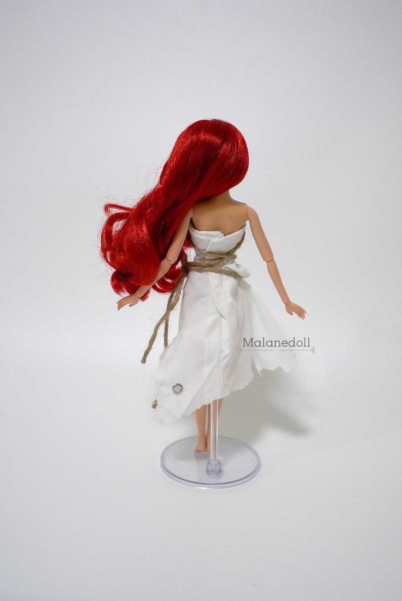 May include: A red-haired doll wearing a white dress with a rope belt. The doll is standing on a clear plastic stand. The text "Malanedoll" is visible on the dress.