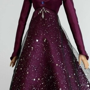 Puede incluir: Un vestido largo de sat&eacute;n morado de manga larga, con cuello de encaje negro y un adorno de cuentas plateadas. El vestido tiene una superposici&oacute;n brillante plateada y una falda de tul negra.