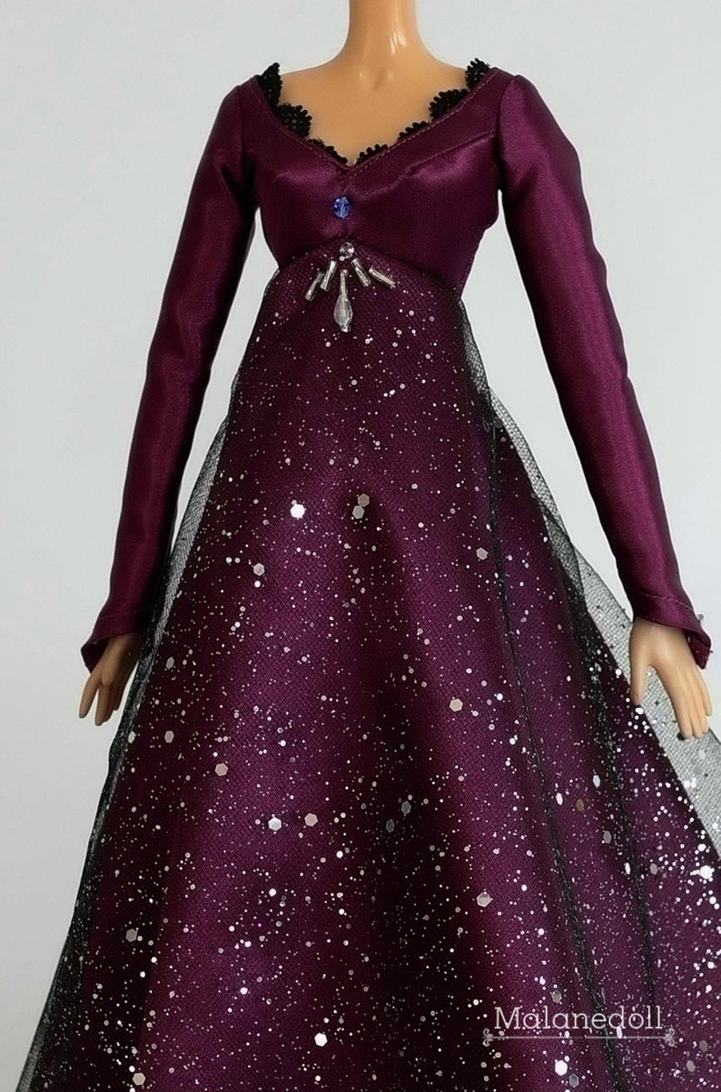 Puede incluir: Un vestido largo de sat&eacute;n morado de manga larga, con cuello de encaje negro y un adorno de cuentas plateadas. El vestido tiene una superposici&oacute;n brillante plateada y una falda de tul negra.