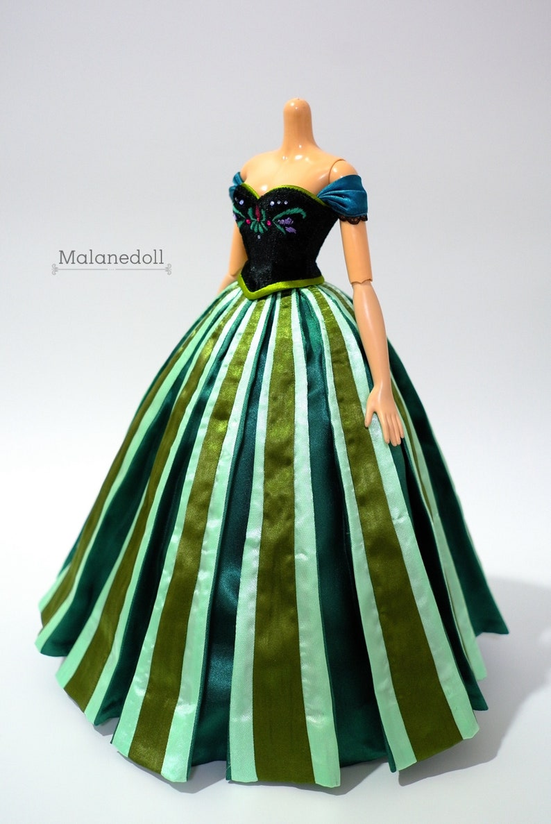 May include: A doll wearing a green and black dress with a striped skirt. The dress has a black bodice with a floral design and a green sash.