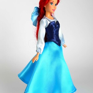 K&ouml;nnte beinhalten: Puppe einer Figur mit langen roten Haaren und einer blauen Schleife. Die Puppe tr&auml;gt ein wei&szlig;es Langarm-Oberteil mit einem dunkelblauen Oberteil und einen langen, hellblauen Satinrock. Schwarze Schuhe vervollst&auml;ndigen den Look. Die Puppe steht auf einem transparenten St&auml;nder.