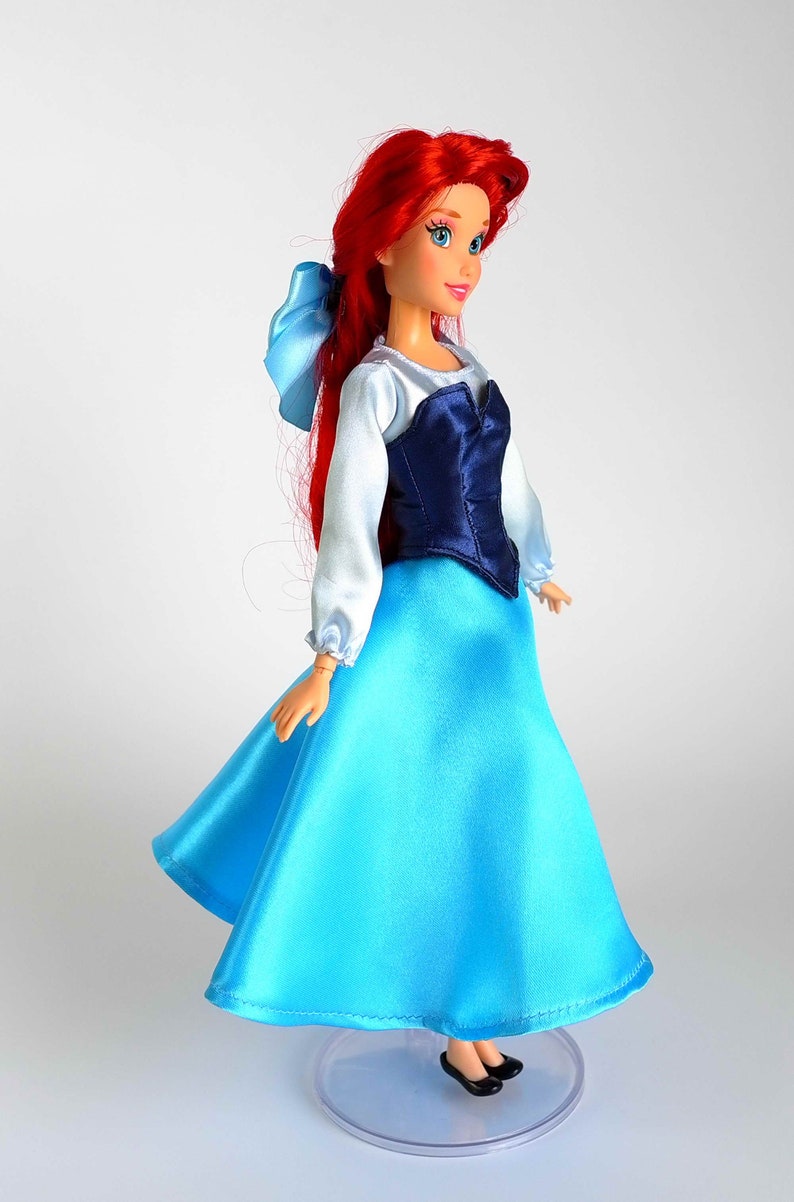 K&ouml;nnte beinhalten: Puppe einer Figur mit langen roten Haaren und einer blauen Schleife. Die Puppe tr&auml;gt ein wei&szlig;es Langarm-Oberteil mit einem dunkelblauen Oberteil und einen langen, hellblauen Satinrock. Schwarze Schuhe vervollst&auml;ndigen den Look. Die Puppe steht auf einem transparenten St&auml;nder.