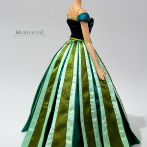 May include: A doll wearing a green and black dress with a striped skirt. The dress has a sweetheart neckline and off-the-shoulder straps.