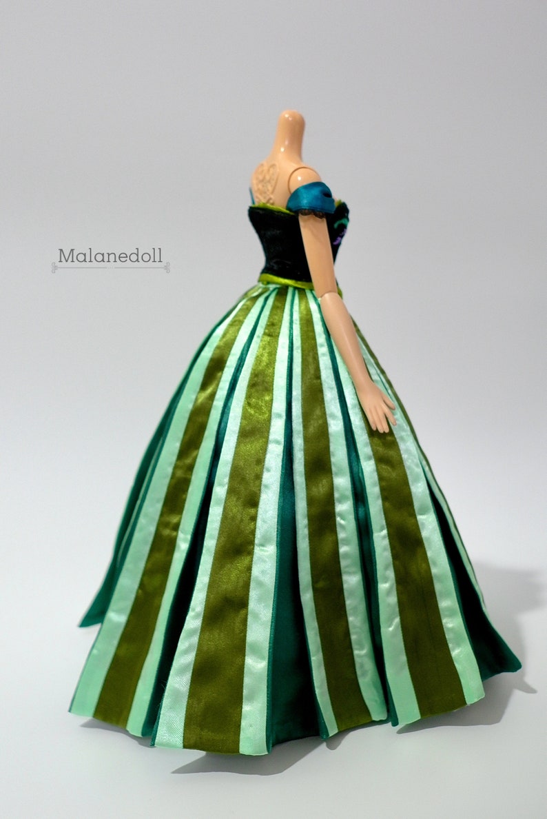 May include: A doll wearing a green and black dress with a striped skirt. The dress has a sweetheart neckline and off-the-shoulder straps.