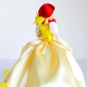 Pode incluir: Uma boneca de cabelo ruivo vestindo um vestido de baile de cetim amarelo com um corpete branco e um la&ccedil;o amarelo no cabelo. O vestido tem uma bainha amarela com babados.