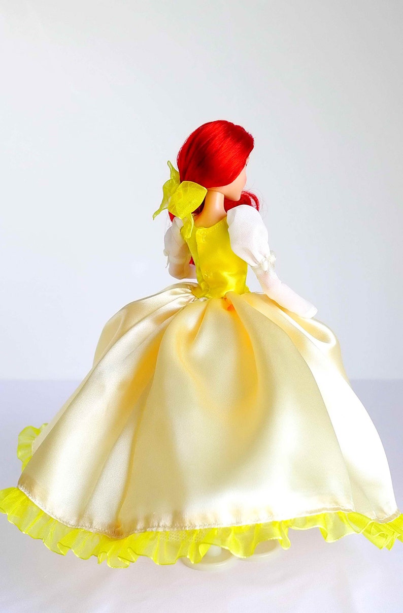 Pode incluir: Uma boneca de cabelo ruivo vestindo um vestido de baile de cetim amarelo com um corpete branco e um la&ccedil;o amarelo no cabelo. O vestido tem uma bainha amarela com babados.
