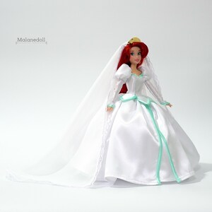 May include: A white bridal gown with a long veil and a green sash. The doll is wearing a gold crown and has red hair.