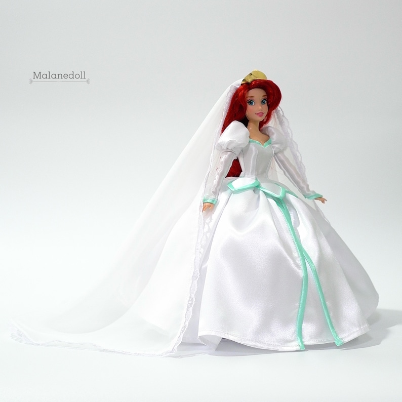 May include: A white bridal gown with a long veil and a green sash. The doll is wearing a gold crown and has red hair.