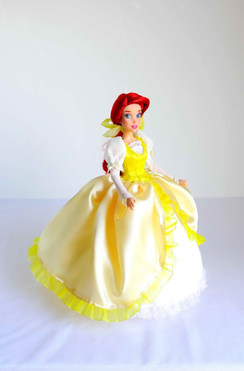 Pode incluir: Uma boneca de cabelo ruivo vestindo um vestido de cetim amarelo com uma borda de renda branca e uma fita amarela no cabelo.