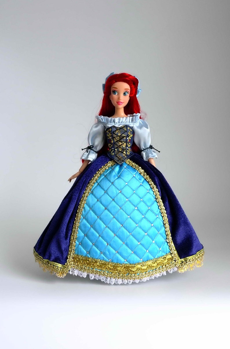 May include: A red-haired doll wearing a blue and gold dress with a white lace trim. The doll has a blue bodice with a gold and black design. The doll is wearing a blue and gold skirt with a white lace trim.