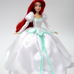 May include: A red-haired doll wearing a white satin ball gown with a light green trim. The doll has blue eyes and a red lip. The gown has a sweetheart neckline and a full skirt.