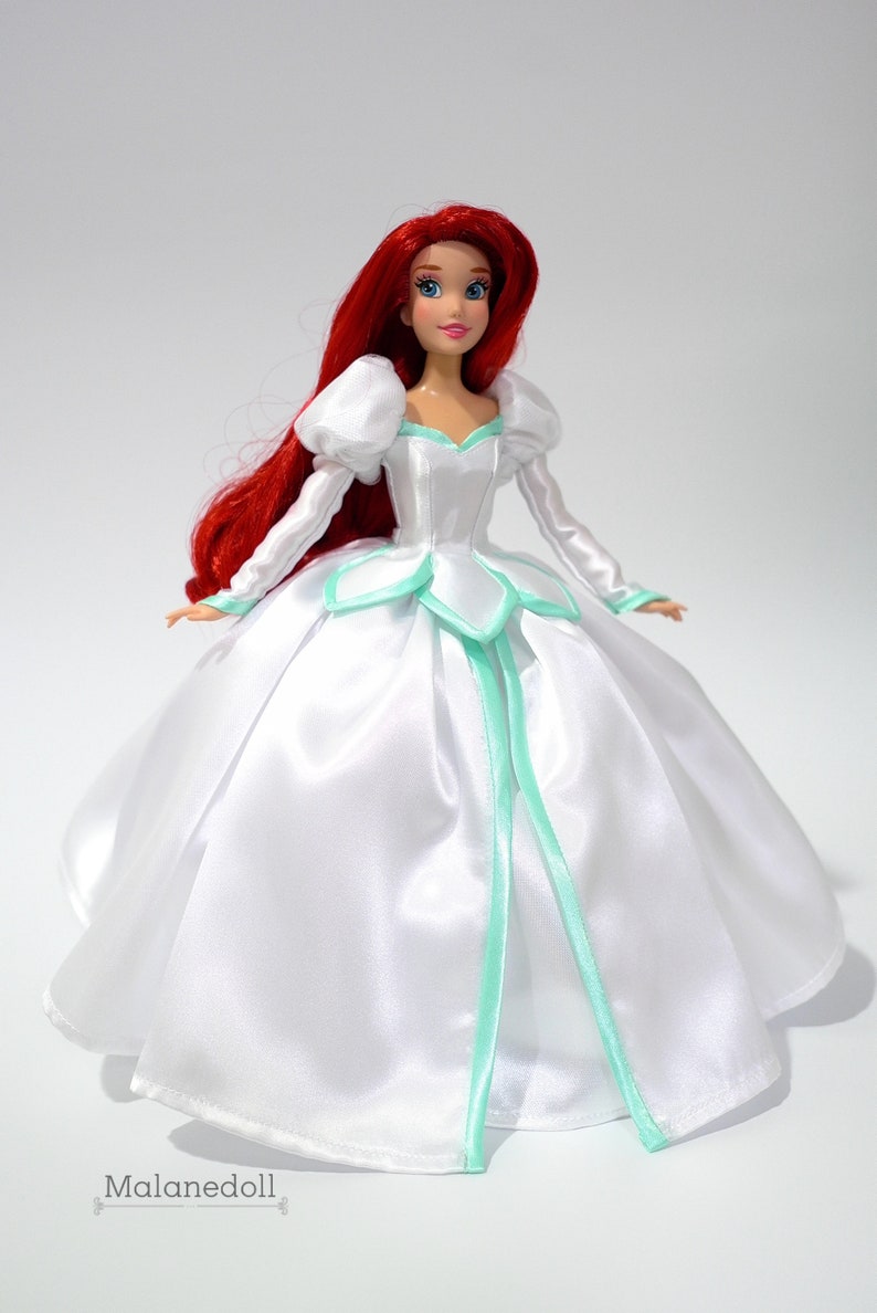 May include: A red-haired doll wearing a white satin ball gown with a light green trim. The doll has blue eyes and a red lip. The gown has a sweetheart neckline and a full skirt.