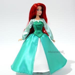 May include: A red-haired doll wearing a green and teal satin dress with a white underskirt. The doll has blue eyes and is wearing a green bodice. The doll is standing on a clear stand. The text "Malanedoll" is visible in the lower right corner of the image.
