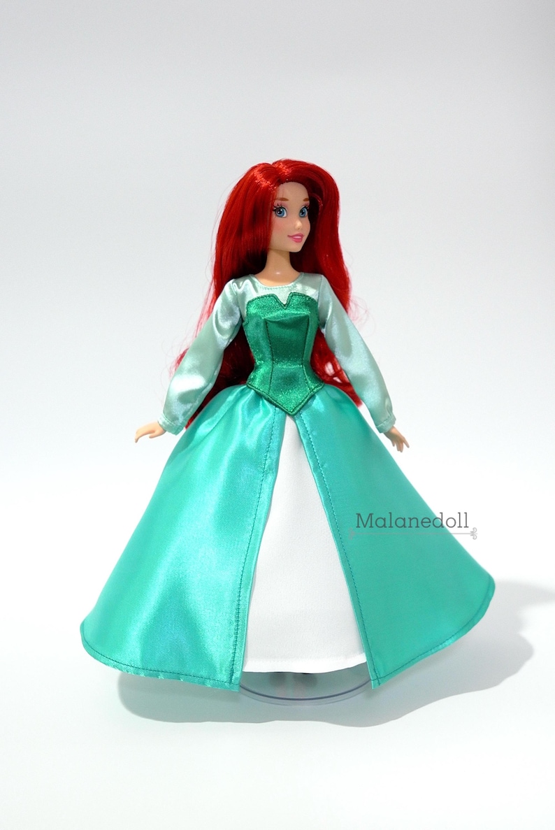 May include: A red-haired doll wearing a green and teal satin dress with a white underskirt. The doll has blue eyes and is wearing a green bodice. The doll is standing on a clear stand. The text "Malanedoll" is visible in the lower right corner of the image.