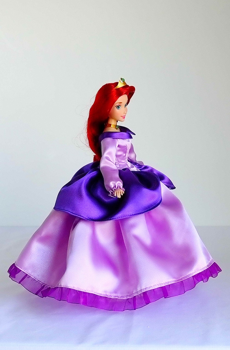 May include: A red-haired doll wearing a purple and pink dress with a ruffled hem. The doll is wearing a gold crown with green gems.