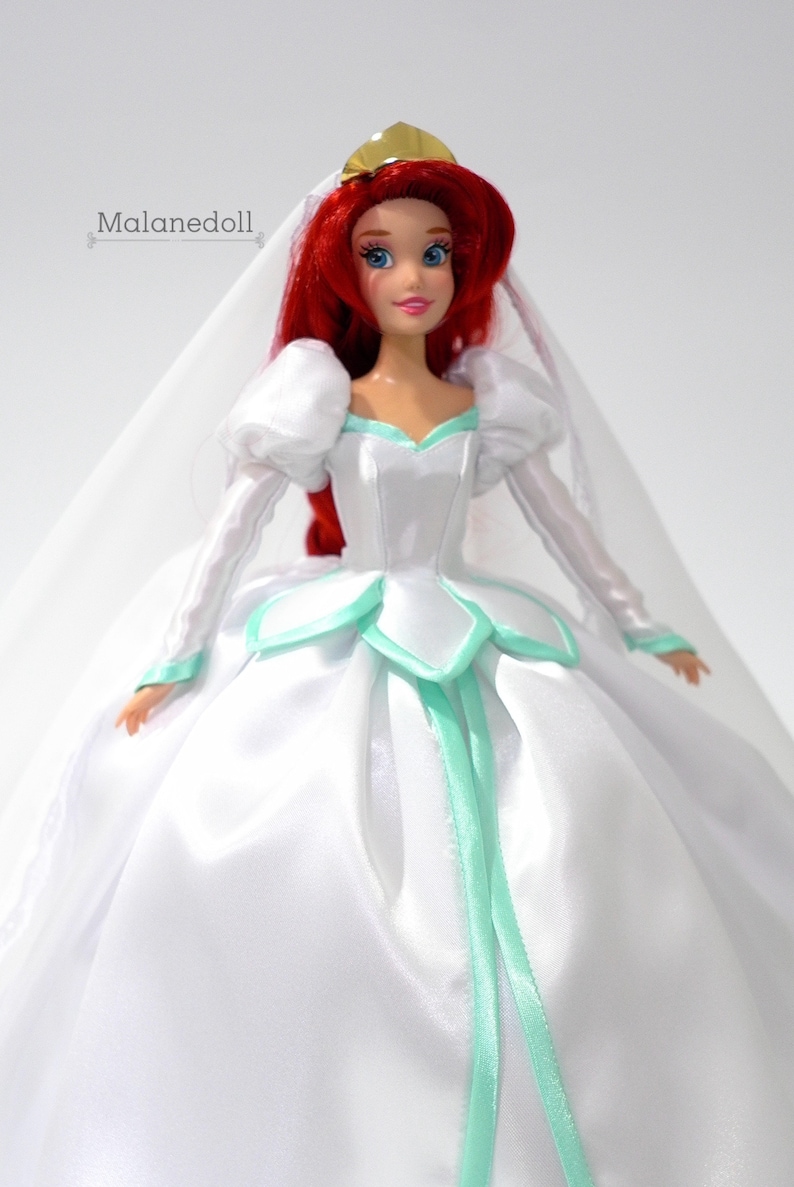 May include: A red-haired doll wearing a white wedding dress with a green sash and a veil. The doll is wearing a gold crown.