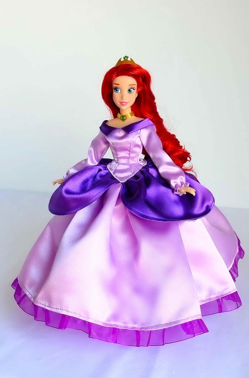 May include: A red-haired doll wearing a purple and pink satin ball gown with a green gem necklace and a gold crown.