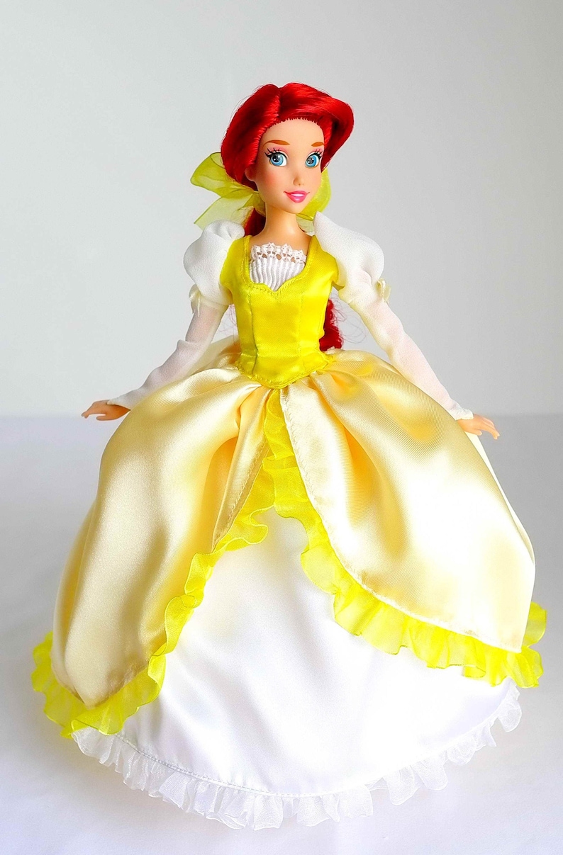 Pode incluir: Uma boneca de cabelo ruivo vestindo um vestido de cetim amarelo com renda branca e um la&ccedil;o amarelo no cabelo.